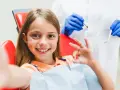 Uśmiechnięta dziewczynka po zabiegu lakierowania zębów u dzieci pokazuje znak "OK".