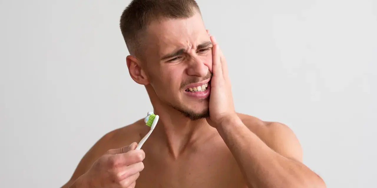 Mężczyzna z bólem zęba, trzymający szczoteczkę do zębów. Jego twarz wykrzywiona jest w grymasie, a ręka przyciśnięta do policzka, co sugeruje, że ma opuchnięte dziąsła.