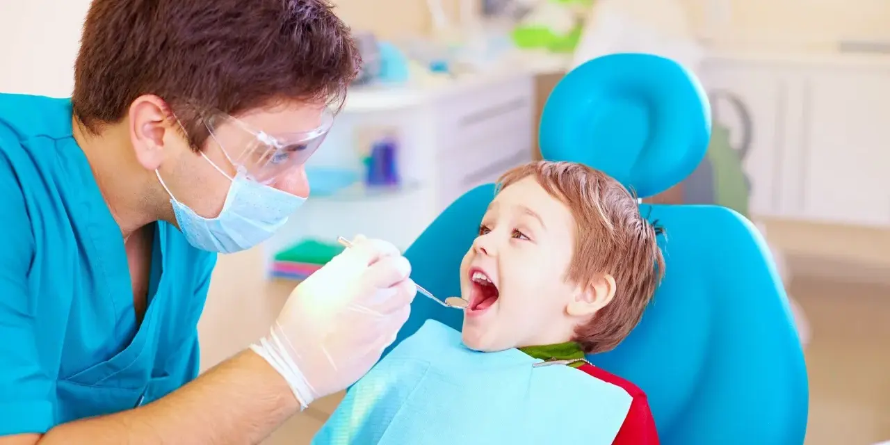 Dentysta bada uśmiechniętego chłopca, sprawdzając, kiedy najpóźniej pierwszy ząbek pojawi się u malucha.
