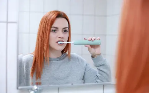 Rudowłosa kobieta myje zęby szczoteczką soniczną. Czy szczoteczka soniczna jest dobra? Ta kobieta wydaje się zadowolona.