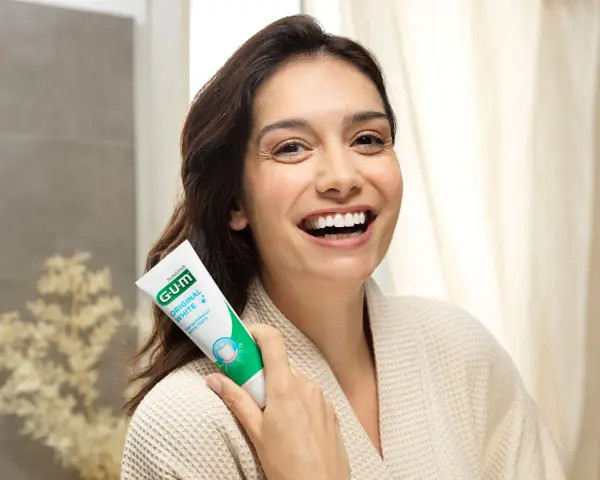 Uśmiechnięta kobieta trzymająca pastę GUM Original White. Dbanie o dziąsła (gum care) to klucz do pięknego uśmiechu.