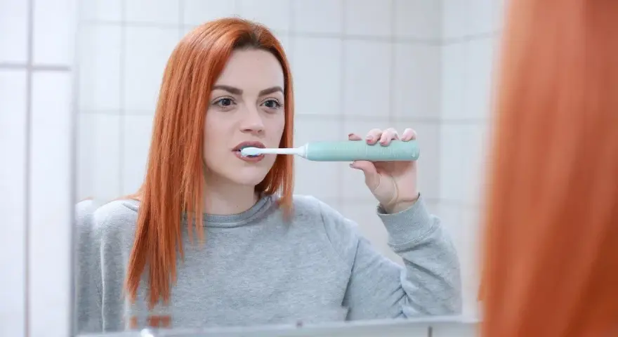 Rudowłosa kobieta myje zęby szczoteczką soniczną, zastanawiając się, czy szczoteczka soniczna usuwa kamień.