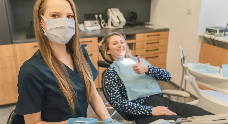 Uśmiechnięta pacjentka daje kciuk w górę podczas pierwszej wizyty u dentysty po latach. Dentystka w masce i rękawiczkach przygotowuje narzędzia.
