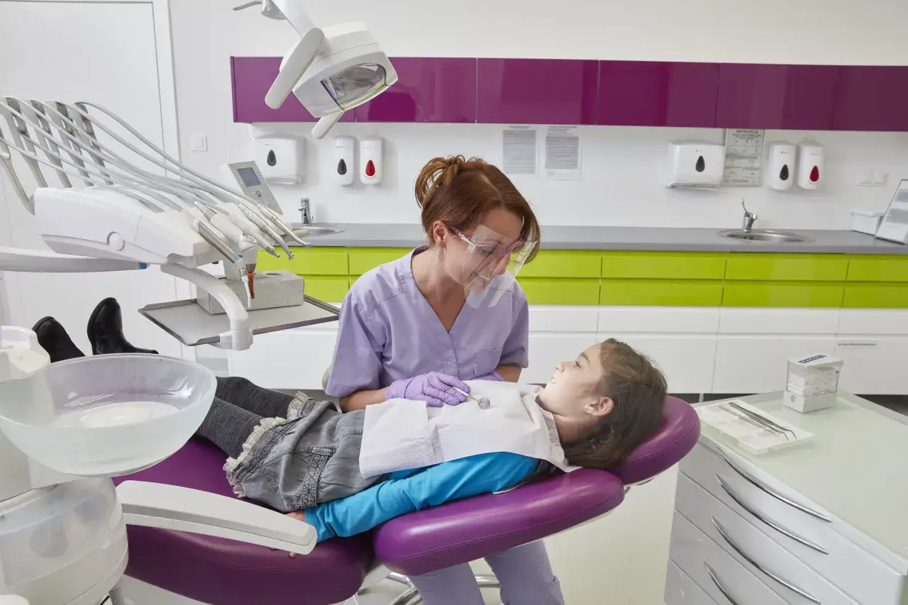Dentystka bada uśmiechniętą dziewczynkę, która leży na fotelu. Czy to już czas, kiedy najpóźniej pierwszy ząbek?