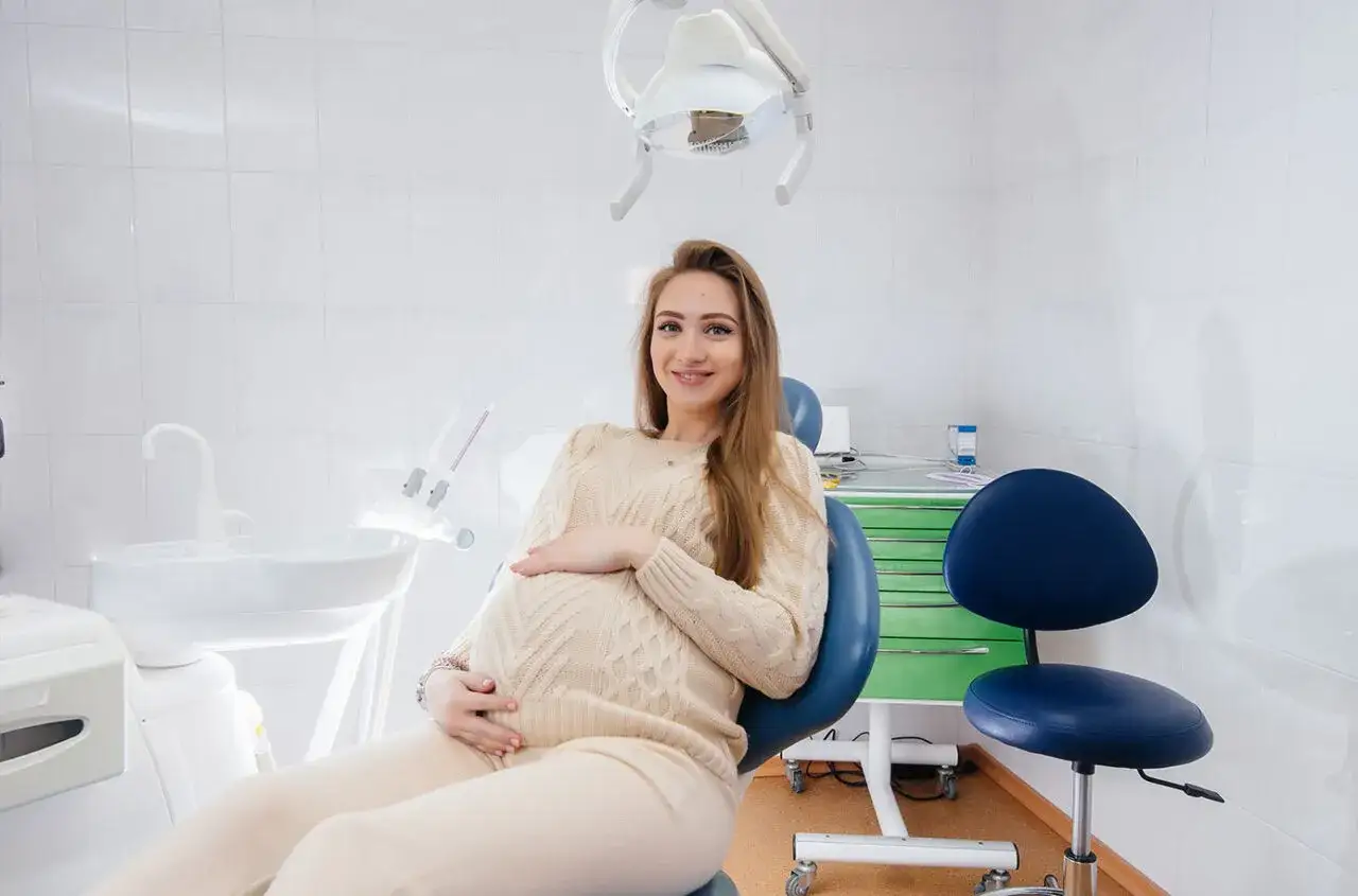 Uśmiechnięta ciężarna kobieta w gabinecie stomatologicznym, przygotowująca się do fluoryzacji. Dba o zdrowie zębów dla siebie i dziecka.