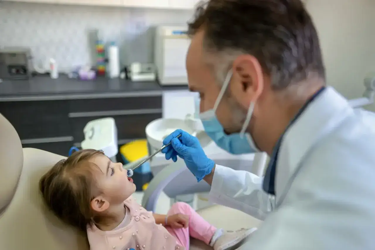 Dentysta bada uśmiech małej dziewczynki, sprawdzając, kiedy najpóźniej pierwszy ząbek pojawi się w jej buzi.