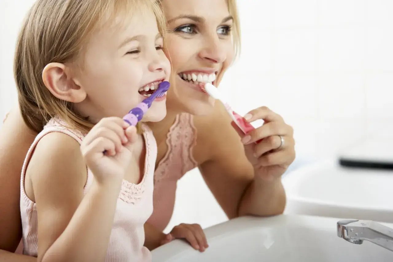 Mama i córka myją zęby. Uśmiechnięta dziewczynka z fioletową szczoteczką, mama z różową. Wesoła instrukcja mycia zębów dla dzieci.